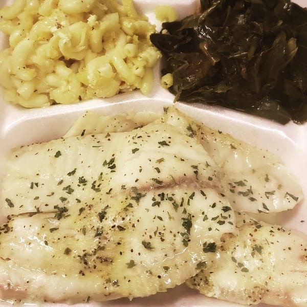 Southern soul food meal with baked fish, macaroni and cheese, and collard greens in a to-go container.